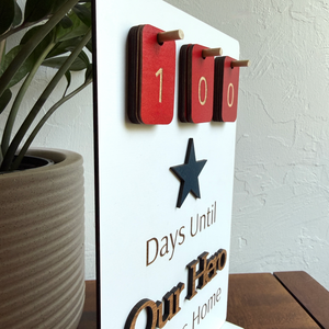 Decorative sign with '100 Days Until Our Hero Home' text and wooden numbers on a white background.