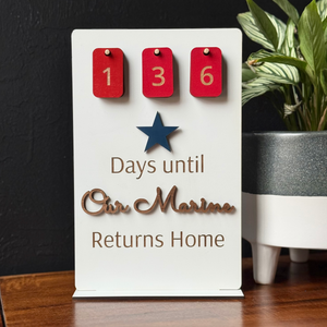 Decorative sign with red tags and a star, counting days until a marine returns home, on a wooden surface.