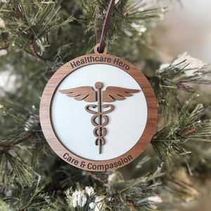Wooden ornament with caduceus symbol and 'Healthcare Hero Care & Compassion' text on a Christmas tree.