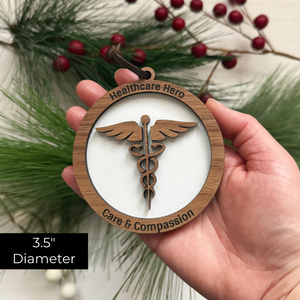 Hand holding a wooden ornament with 'Healthcare Hero' and 'Care & Compassion' text, surrounded by greenery and berries.
